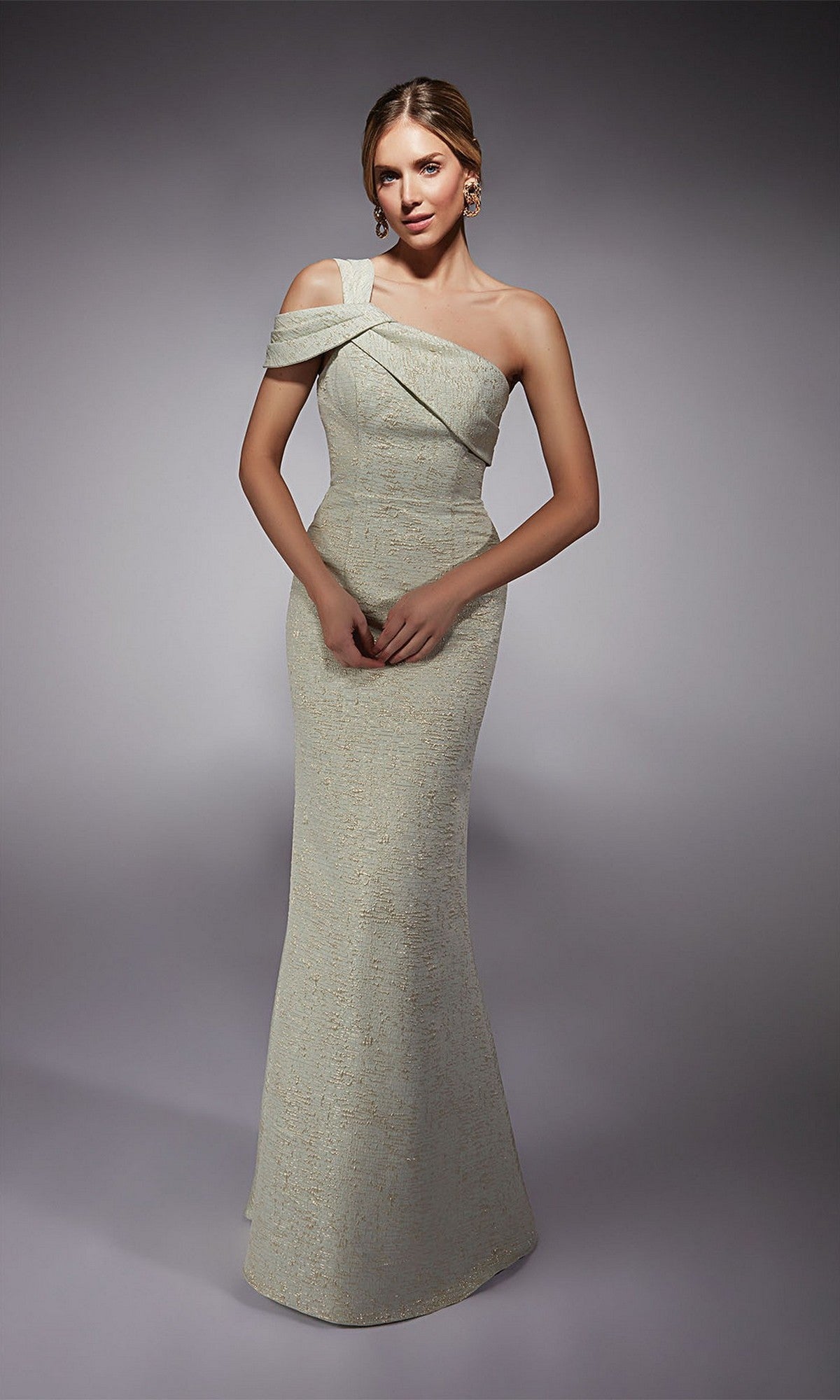 A woman wears the Alyce 27749 long formal dress in sea foam/gold, featuring a fitted sheath silhouette, floor-length design, cold-shoulder sleeves, and a subtle shimmer, set against a plain gray background.